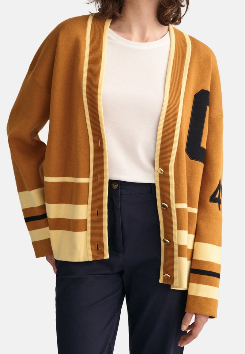 Femme portant un cardigan marron avec des rayures beiges et des détails noirs numérotés, une chemise blanche et un pantalon foncé sur fond blanc.