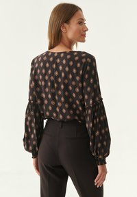 Dunkel gemusterte Bluse mit Diamantformen in Brauntönen, mit Puffärmeln und lässiger Passform, kombiniert mit körperbetonten schwarzen Hosen.
