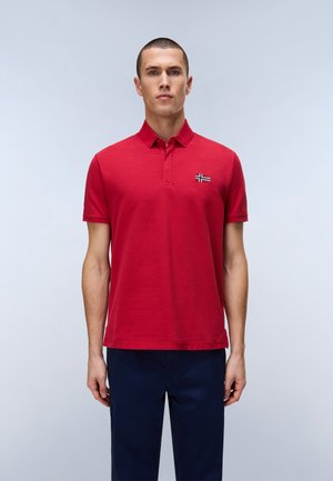 Giovane uomo con capelli corti in piedi, indossa una polo rossa a maniche corte liscia e pantaloni blu scuro, davanti a uno sfondo grigio chiaro.