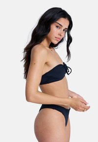 Σετ μαγιό μπικίνι μαύρου χρώματος με τοπάκι bandeau που δένει μπροστά και ψηλόμεσο σλιπ. Λείο ύφασμα με μινιμαλιστικό σχεδιασμό.