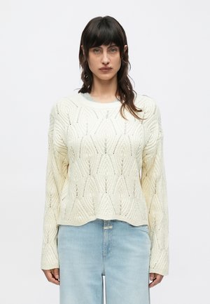 Žena s tmavými vlasy, oblečená v krémovém svetr s copánkovým vzorem a světle modrých džínech s vysokým pasem, stojící proti jednobarevnému bílému pozadí.