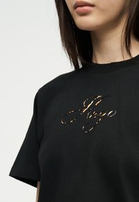 Γυναίκα που φοράει μαύρο μπλουζάκι με το όνομα "Anya" σε καλλιγραφική γραφή, με σχέδιο που συνδυάζει λεοπάρ και μεταλλικό σχέδιο στο στήθος.