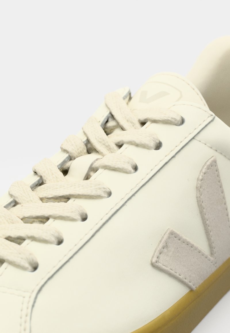 Baskets blanches avec des accents gris clair, lacets plats et une semelle en caoutchouc texturée. Le côté présente un logo proéminent en matériau contrastant.