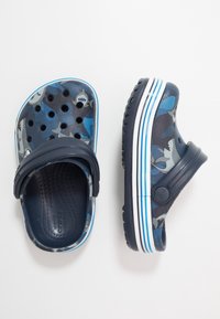 Modré dřeváky s maskáčovým vzorem, ventilačními otvory, silným páskem a bílou pruhovanou podrážkou. Vyrobeny z lehkého materiálu s texturovaným vnitřkem.