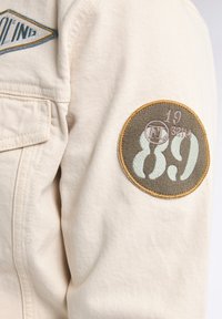 Chaqueta beige con un parche circular verde y marrón que lleva el texto "89" y "19 3214", además de un logo bordado encima del parche.
