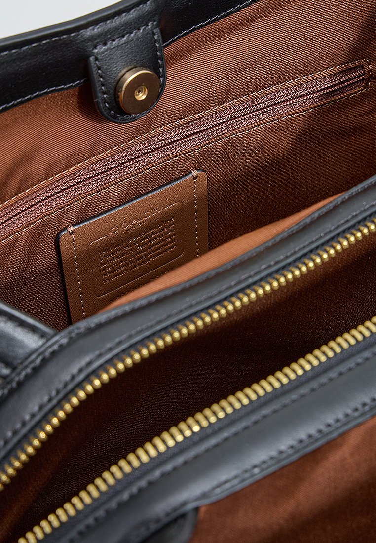 Interno di una borsa a tracolla in pelle nera, con rivestimento in tessuto marrone, zip dorata e un'etichetta con testo in rilievo. Tasca con zip visibile su un lato.