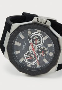 Montre octogonale avec un bracelet en silicone noir texturé, un cadran gris à motifs, des accents en argent, et des aiguilles rouges. Dotée de trois sous-cadrans.