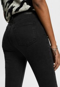Jeans de mezclilla negra con un corte ajustado, que cuentan con bolsillos traseros, una cintura alta y una etiqueta de marca en la cinturilla. Tela texturizada visible.