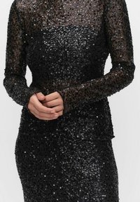 Robe noire en sequins avec manches longues et silhouette ajustée. Le tissu présente un revêtement transparent, orné de nombreux sequins brillants.