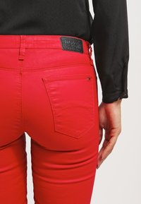 Czerwone dopasowane dżinsy z tylną kieszenią, gładką teksturą oraz czarną skórzaną metką marki przy pasku. Widok z tyłu z czarną bluzką.