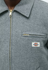 Siva vunena jakna s ovratnikom, zlatnim patentnim zatvaračem i lijevim džepom na prsima. Ima Dickies logo aplikaciju. Teksturu tkanine.