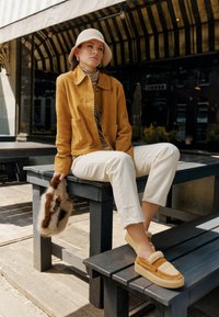 Yellow suede jacket, striped top, white jeans, and brown fur clutch. Beige slip-on shoes with textured accents, seated on a wooden bench.