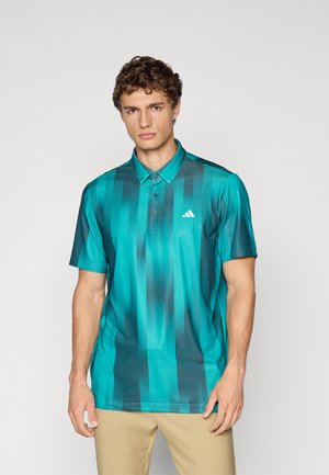 Young man wearing a turquoise and dark teal gradient short-sleeve polo shirt with a white logo on the chest and beige pants.
