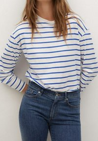 Camisa de manga larga blanca con rayas horizontales azules, combinada con jeans oscuros. Corte casual con cuello redondo y tejido suave.