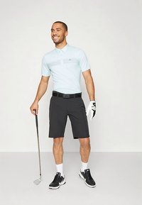 Ανοιχτό μπλε πόλο μπλουζάκι με διακριτό ριγέ σχέδιο, συνδυασμένο με μαύρη βερμούδα. Κρατάει ένα γκολφ, φοράει μαύρη ζώνη και γάντια.