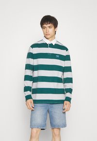 Poloshirt met lange mouwen in groen en grijs horizontale strepen, met een kraag en een klein logo op de linkerborst, gecombineerd met denim shorts.