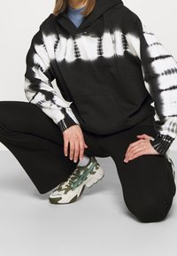 Sudadera a rayas en blanco y negro con bolsillo frontal, combinada con pantalones negros y zapatillas deportivas texturizadas con acentos verdes.