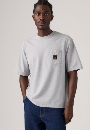 Homme aux cheveux tressés portant un T-shirt gris clair à manches courtes avec une poche poitrine et un jean bleu, debout devant un fond uni.