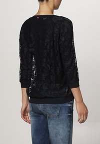 Maglione in pizzo nero con maniche a 3/4, dotato di orlo e polsini neri solidi. Tessuto in pizzo trasparente con motivi floreali, indossato sopra jeans blu.