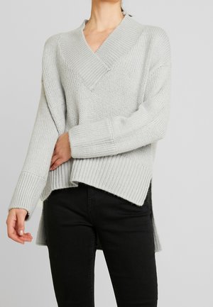 Kobieta w jasnoszarym, dzianinowym swetrze z dekoltem w serek i długimi rękawami oraz czarnych spodniach, stojąca na jednolitym tle.