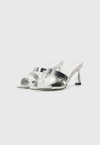 Zilveren metallic hoge hak mules met open teen en kruisbanden, voorzien van een decoratieve accent en een slanke hak met een zwarte punt.