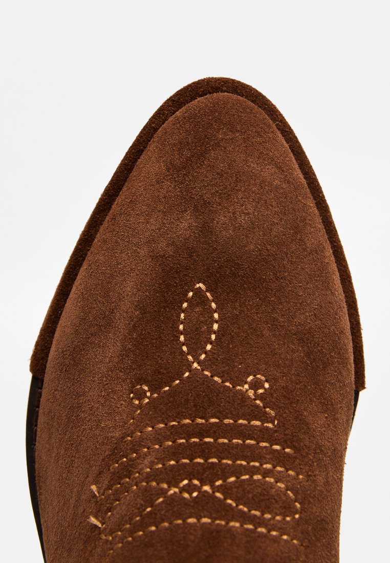 Braune Wildleder-Cowboy-Stiefel mit spitzer Zehe, ausgestattet mit beigem Nähen in einem wellenförmigen Muster nahe der Zehenpartie. Glatte Textur durchgehend.