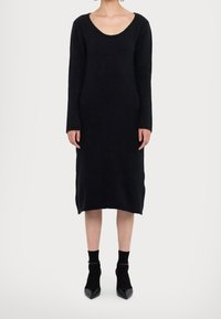 Femme portant une robe midi en maille noire à manches longues, associée à des chaussettes noires jusqu'à la cheville et des escarpins noirs à bout pointu sur fond blanc.
