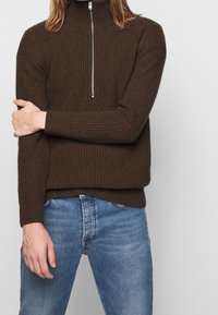 Brązowy sweter z prążkowanej dzianiny z wysokim zamkiem, z długimi rękawami i luźnym krojem, w zestawieniu z jasnoniebieskimi dżinsami.