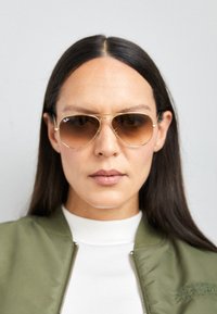 Ochelari de soare Ray-Ban aviator cu ramă din metal aurie și lentile maro cu degradé, având o bară subțire de sprânceană și logo pe lentilă.