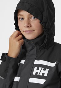 Černá voděodolná bunda s přiléhavou kapucí, která má bílé horizontální pruhy a logo "HH". Látka má hladkou strukturu s matným povrchem.