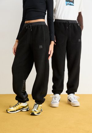 Schwarze Fleece-Jogginghosen mit elastischem Bund und Taschen. Kombiniert mit sportlichen Schuhen; ein Paar hat gelbe Akzente, das andere in Grau und Weiß.