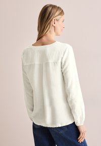 Langærmet cremebluse med tekstureret ryg, rund halsudskæring og afslappet pasform, parret med mørke jeans, der fremhæver en afslappet stil.