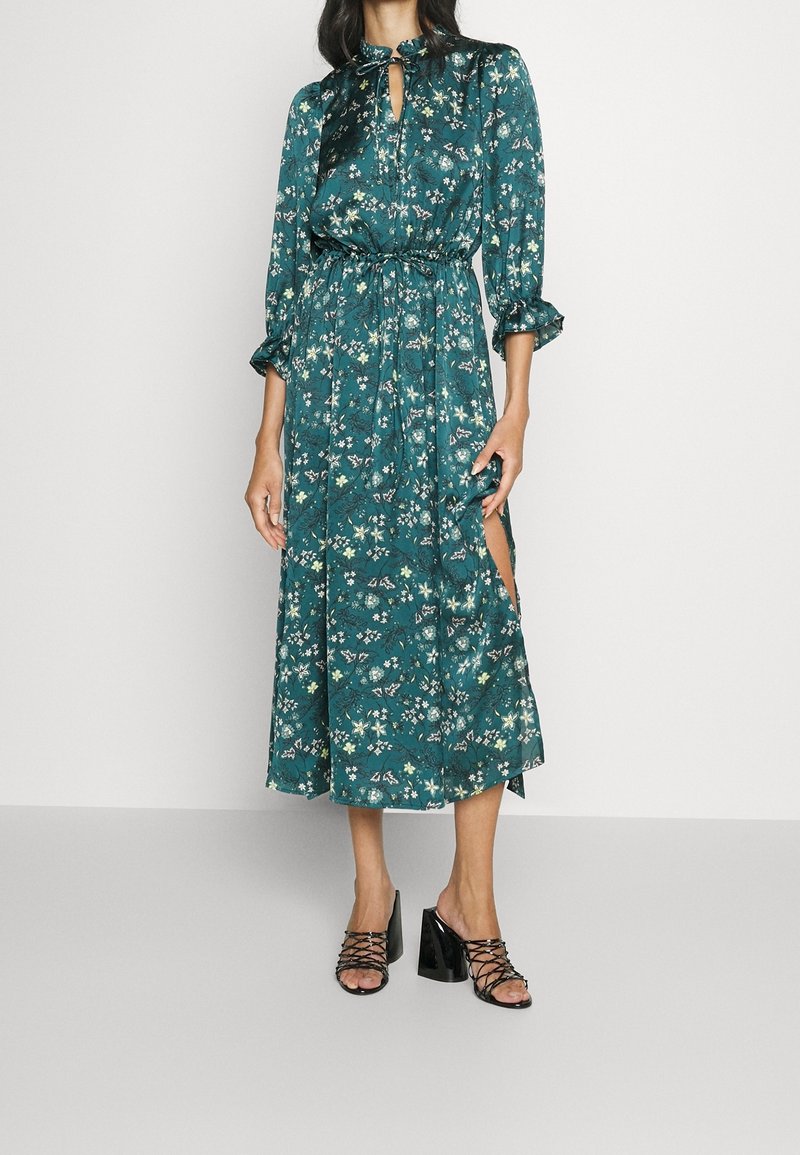 Largo vestido teal con patrón floral, escote en forma de llave y cintura fruncida; cuenta con mangas largas y abertura lateral. Combinado con sandalias de tacón negras.