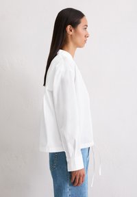 White, long-sleeve blouse with a relaxed fit, rounded collar, side ties, and buttoned cuffs, paired with light blue denim jeans.