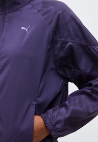 Κλείσιμο λήψης σε άτομο που φοράει μωβ μπουφάν Puma με φερμουάρ και ελαστικό μανίκι, χέρι στην πλευρική τσέπη, εμφανές λογότυπο Puma στο στήθος.