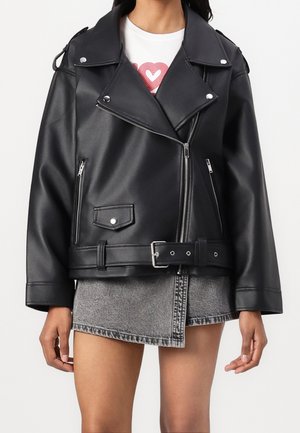 Vrouw draagt een zwarte leren motorjas met zilveren ritsen en een riem, gecombineerd met een grijze denim rok en een wit T-shirt met een rood hart.