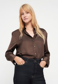 Camisa de satén marrón con botones, cuello y mangas largas, combinada con vaqueros de mezclilla azul oscuro con costuras amarillas y bolsillos delanteros.