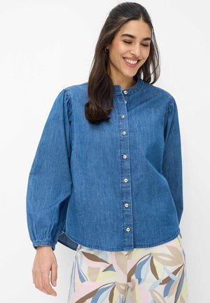 Femme souriante aux longs cheveux foncés portant une chemise en denim bleu boutonnée et un pantalon à motifs aux couleurs pastel, sur un fond blanc.