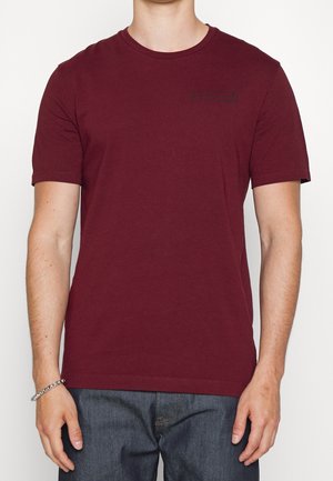 Weinrotes kurzärmeliges T-Shirt aus Baumwolle, mit rundem Ausschnitt und kleinem schwarzem Logo auf der Brust, kombiniert mit blauen Jeans.