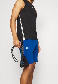 Mustaton urheilu paita valkoisilla sivu raidalla, yhdistettyinä sinisiin shortseihin, joissa on logo ja kuvioitu reunus. Pidessä vastuskuminauhoja.