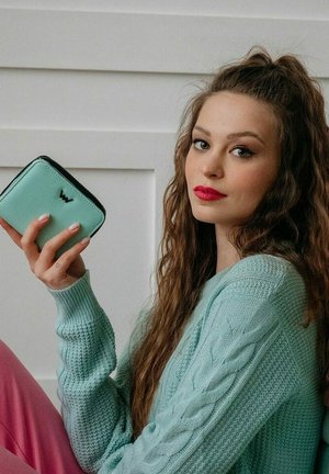 Pistolově zelená kožená peněženka s černým zipem, s logem uprostřed, držená v ruce na pozadí texturovaného svetru.
