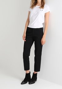 T-shirt en coton blanc avec des manches retroussées et un discret motif graphique sur la poitrine gauche, associé à un pantalon noir ajusté de cheville et des bottines.
