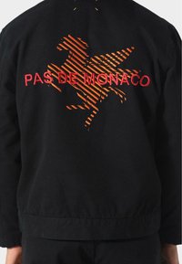 Schwarze Jacke mit rohem Stickerei "PAS DE MONACO". Verfügt über ein abstraktes, oranges Design, eine durchgehende Textur und einen gerippten Saum.