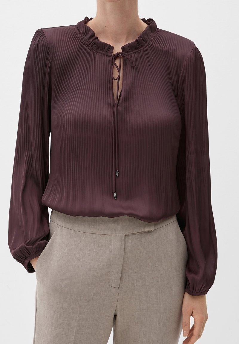 Blouse plissée couleur prune avec un col volanté, des manches longues et un détail en forme de goutte d'eau, associée à un pantalon tailleur crème.