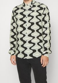Chemise à manches longues boutonnée en vert clair avec un motif ondulé noir et des ovales crème. Dotée d'un col et de boutons noirs arrondis.