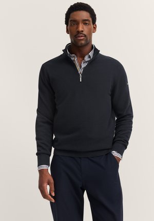 Mann in einem schwarzen Pullover mit Viertelreißverschluss über einem gestreiften Hemd und dunkelblauen Hosen, steht mit einer Hand in der Tasche vor einem hellen Hintergrund.