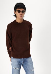 Brun, strikket sweater med rund hals og ribbede manchetter, parret med slidte lyseblå jeans og mørke solbriller.