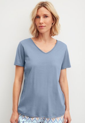Femme blonde portant un t-shirt à manches courtes bleu clair avec col en V et un pantalon à motifs, debout devant un fond blanc uni.