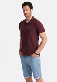 Viininpunainen polo-paita lyhyillä hihoilla ja kauluksella, jossa on pieni logo. Yhdistetty vaaleansinihin farkkushortseihin, rento istuvuus ja käännetyt hihansuut.