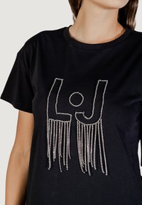 Camiseta de algodón negra con un diseño embellecido con cristales de las letras "LJ" y detalles de flecos colgantes en la parte inferior.